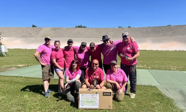 Eine Gruppe von neun Personen in pinken Shirts posiert fröhlich auf einer Rasenfläche vor einer Betonmauer. Eine Frau sitzt vorn und hält ein Schild mit kleinem Text.